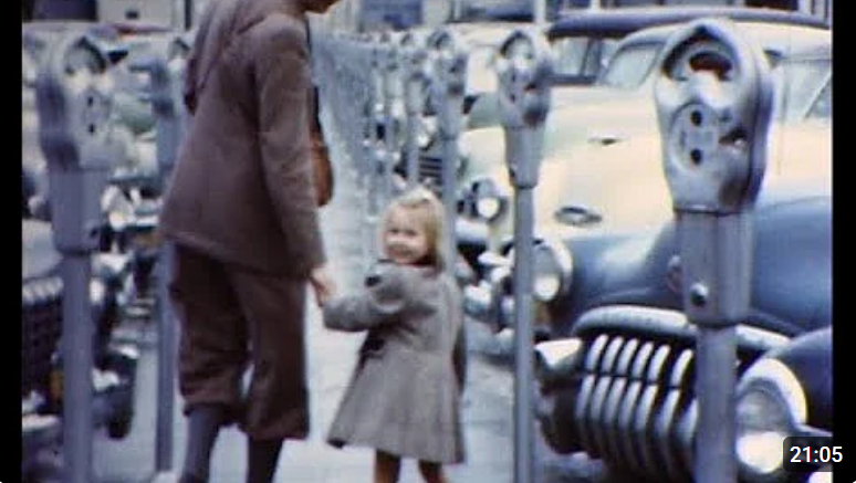 En liten flicka och hennes pappa bland rader av parkerade bilar från början av femtiotalet.